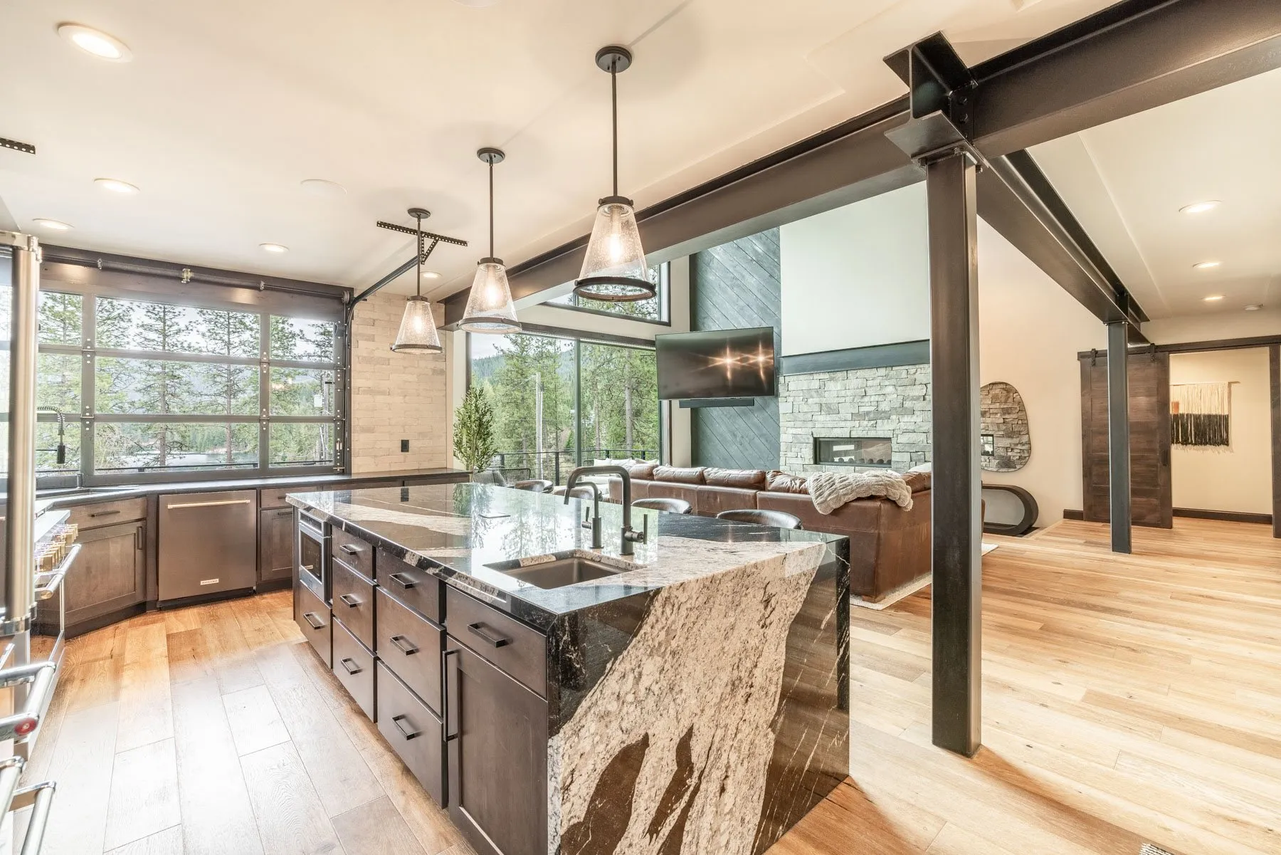 Chef's kitchen with dark cabinetry, a granite waterfall island, pendant lights and black-framed windows to the trees.