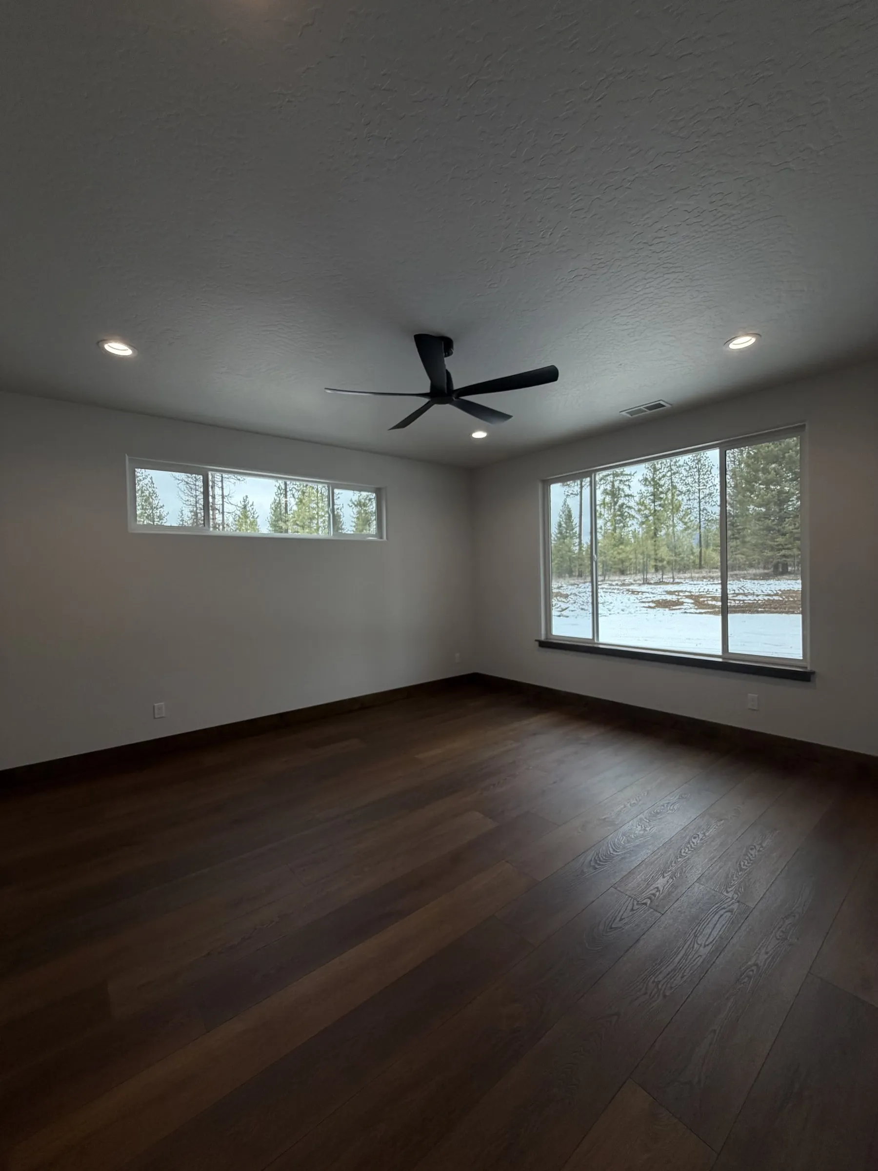 Empty bedroom with dark plank flooring, a black ceiling fan, a clerestory window and a large window to the snowy yard.