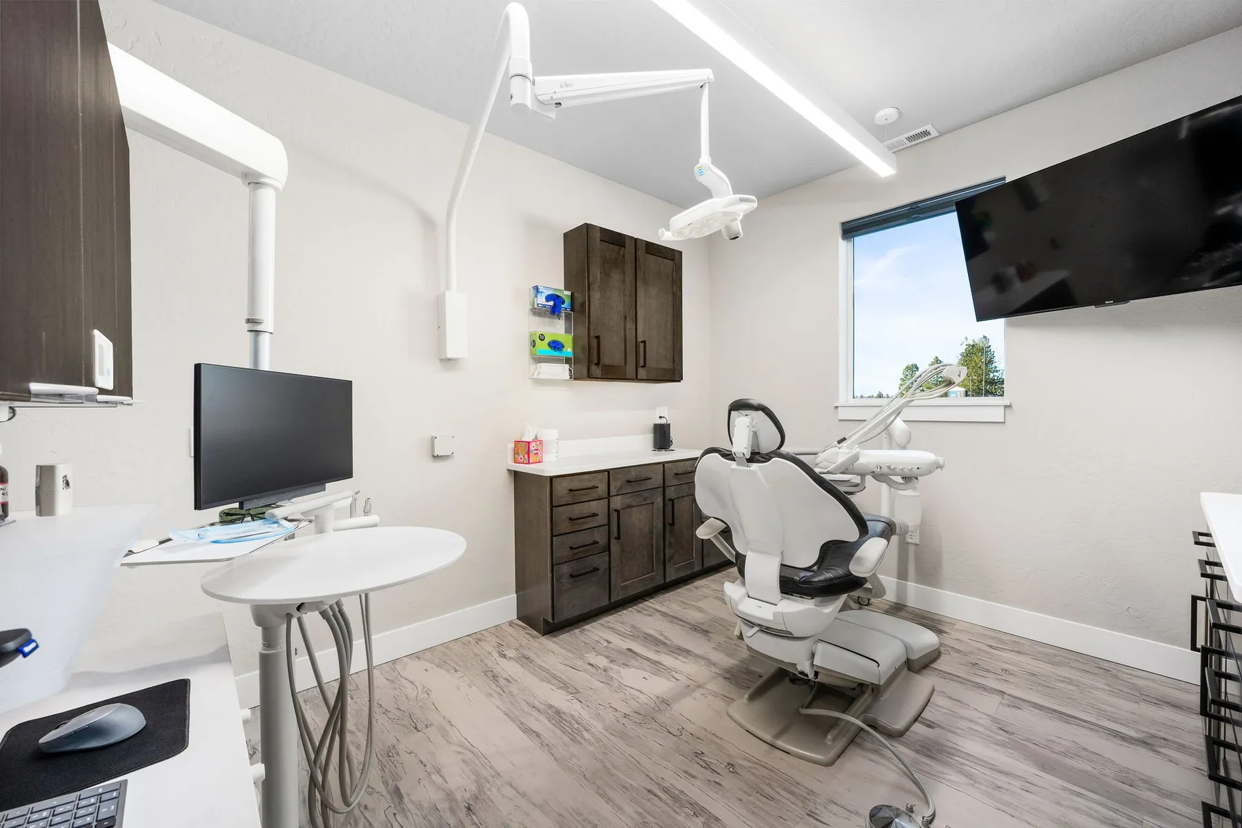 Modern treatment room with a dental chair, overhead light, wall-mounted TV and dark wood cabinetry.