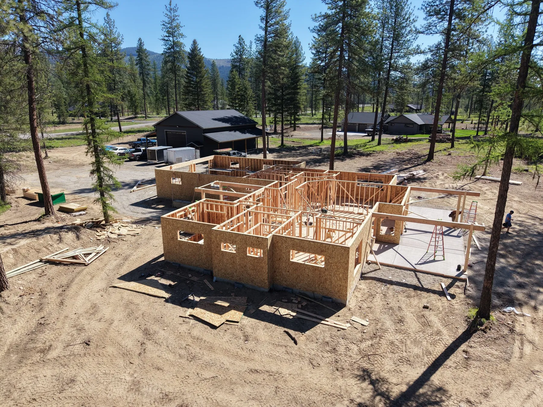 Aerial view of the home being framed on its slab, with the dark shop and neighboring homes behind in the pines.