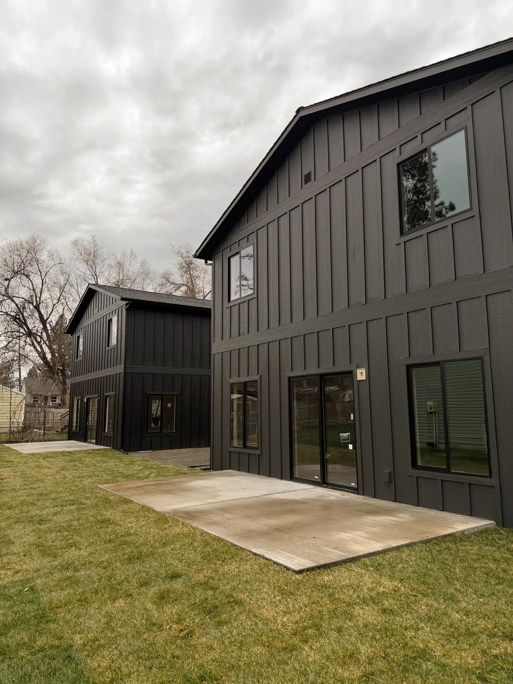 Rear of the twin townhomes, two dark board-and-batten buildings with sliding patio doors opening onto concrete pads and a new lawn.