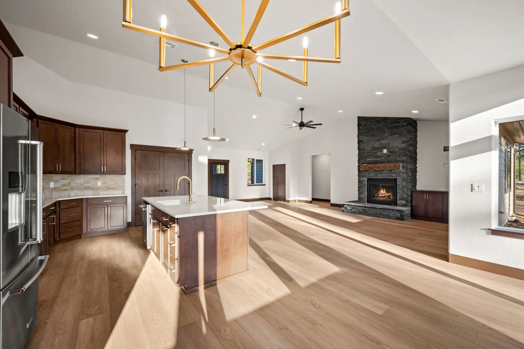 Open kitchen with dark wood cabinetry, a quartz island under a gold sputnik chandelier, and a stacked-stone fireplace in the great room beyond.