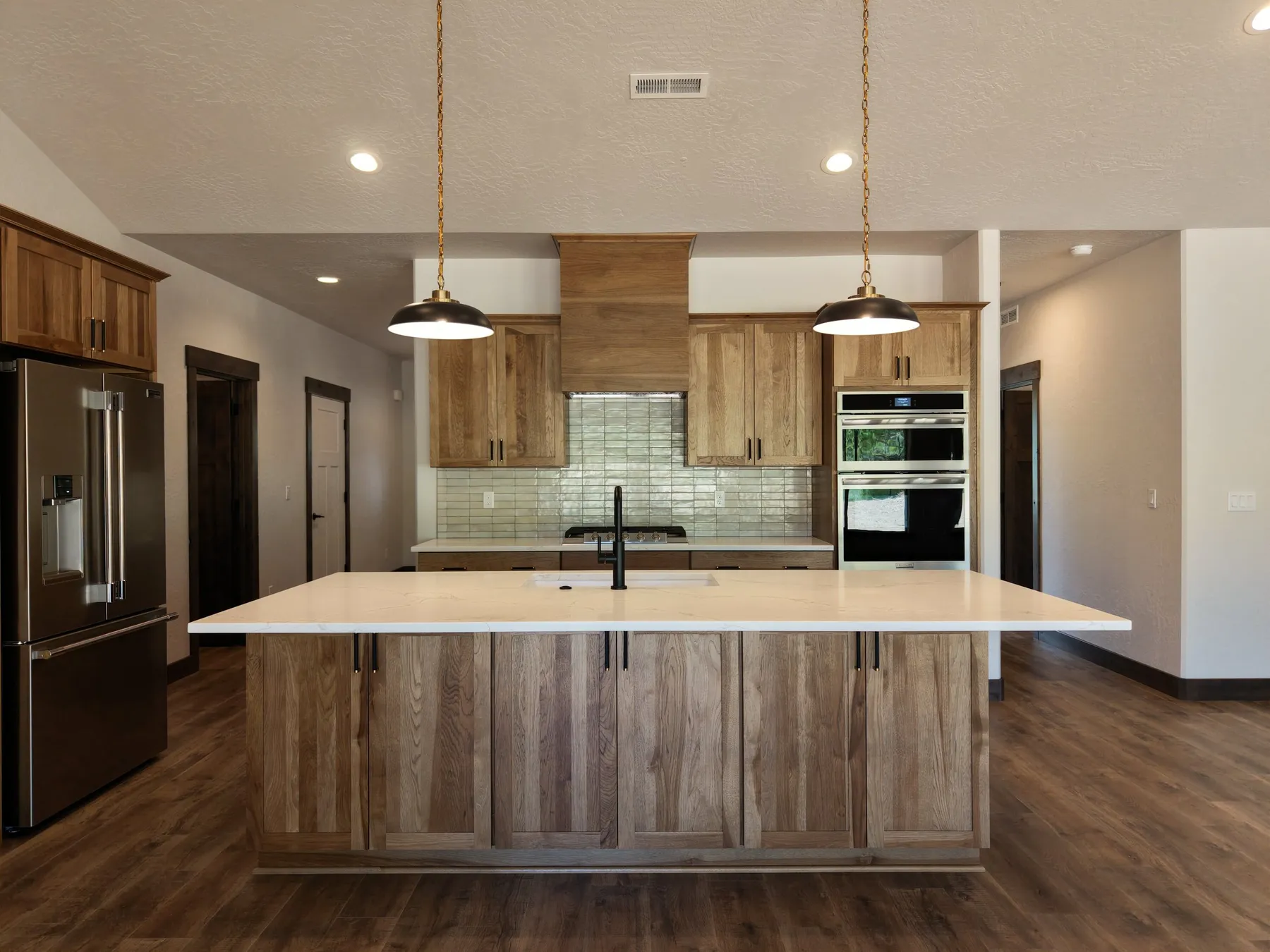 Kitchen with hickory cabinetry, a large white stone island, a tile backsplash, double wall ovens and brass pendant lights.