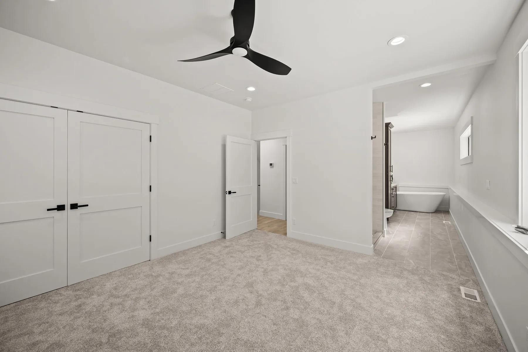 Carpeted bedroom with white double closet doors, a black ceiling fan and an en suite bath with a soaking tub.