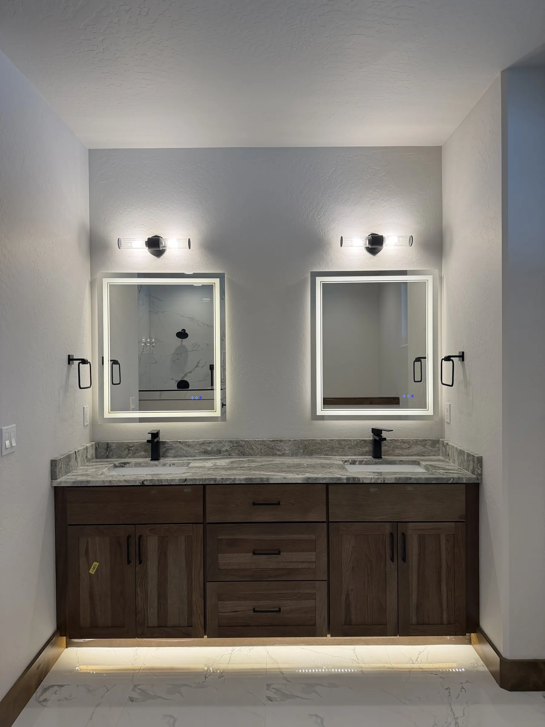 Primary bathroom double vanity with a granite countertop, dark wood cabinets and two backlit mirrors.