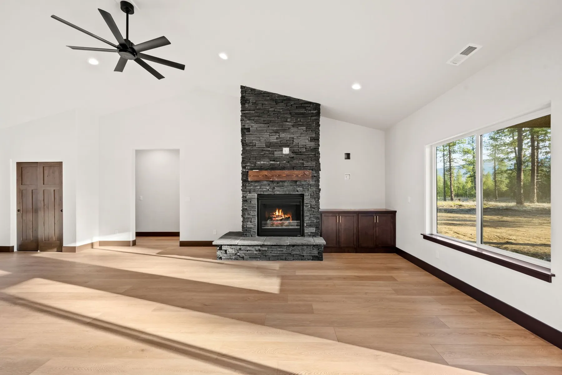 Vaulted great room with a floor-to-ceiling stacked-stone fireplace, wood mantel, light plank flooring and a large window onto the pines.
