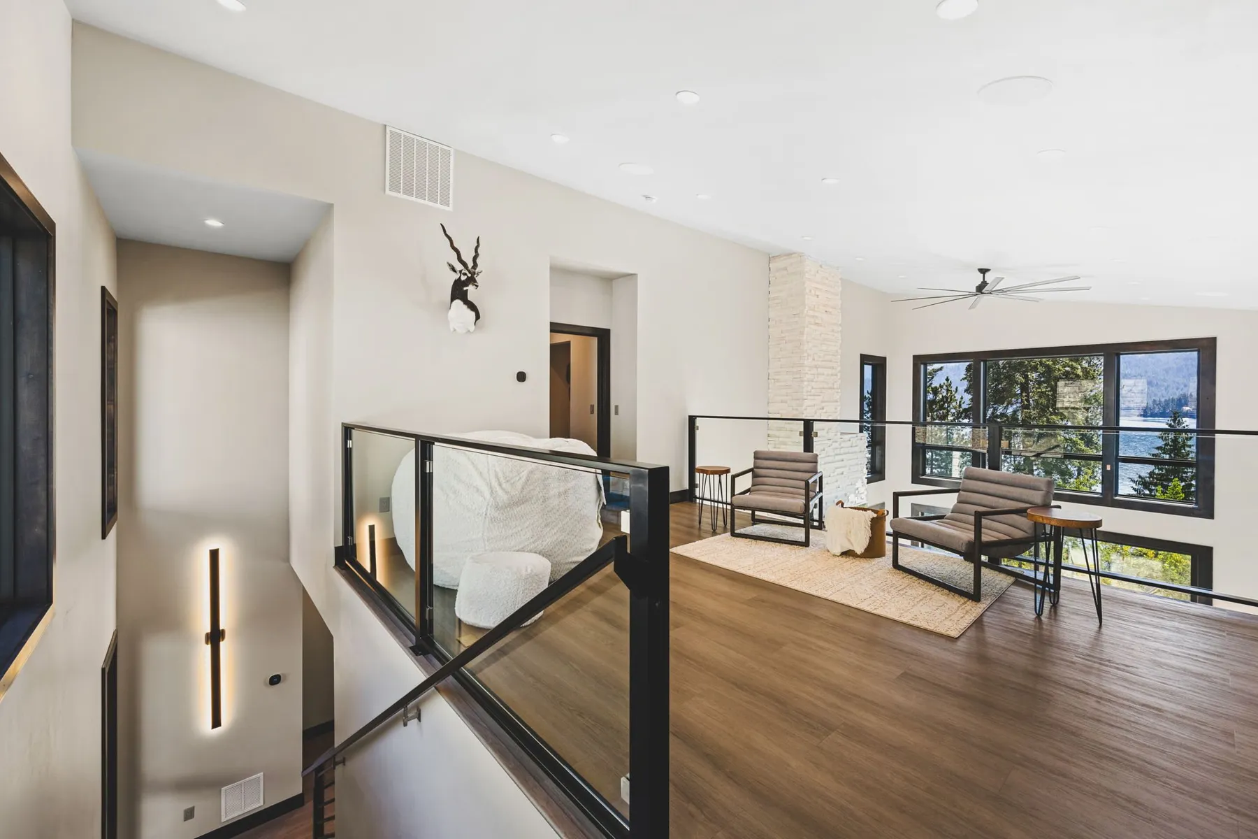 Upper loft with a glass railing overlooking the stairwell, a sitting area and lake views through tall windows.