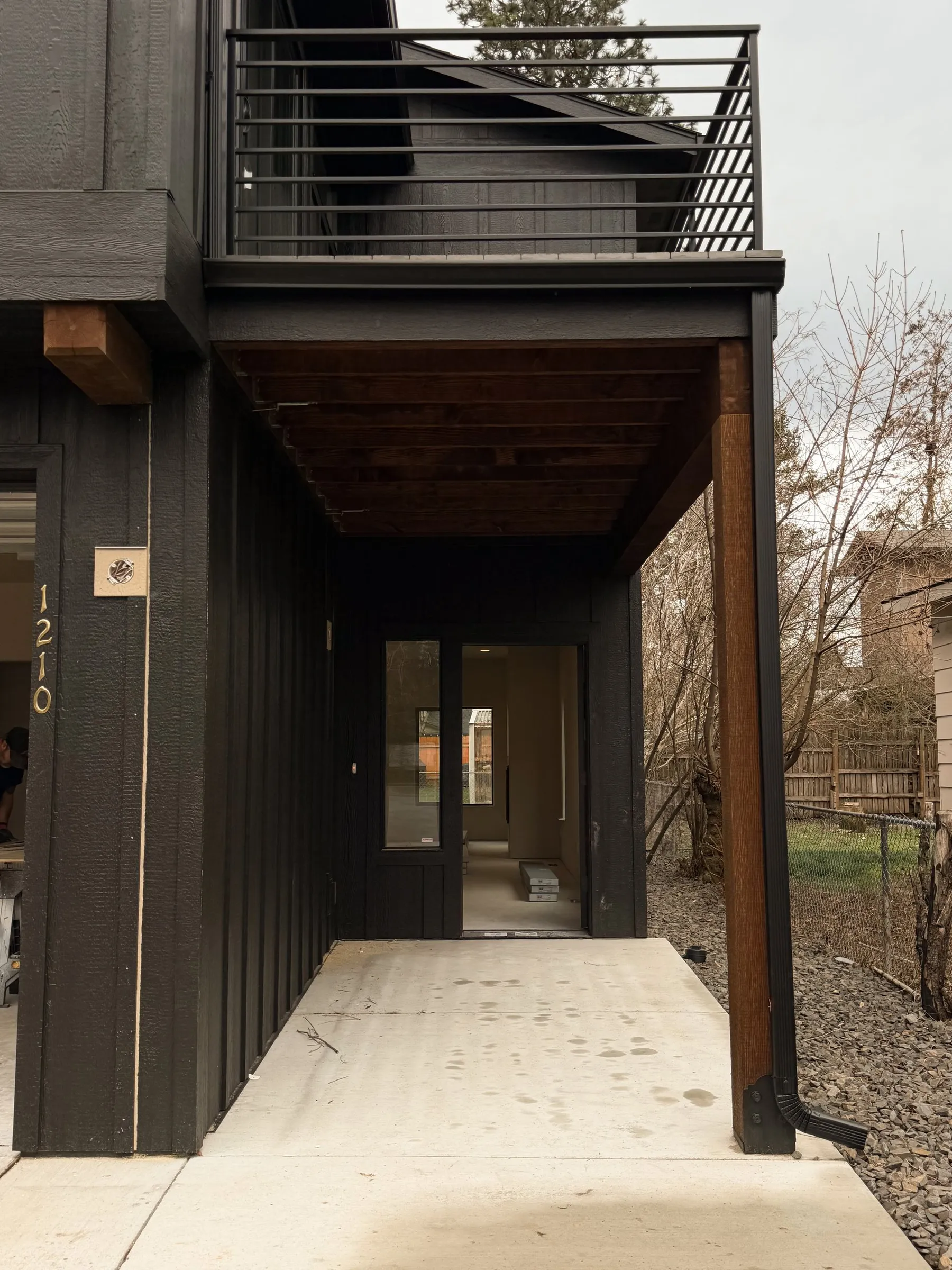 Covered front entry of unit 1210 with a timber post, wood-plank soffit, glass door and the balcony railing above.