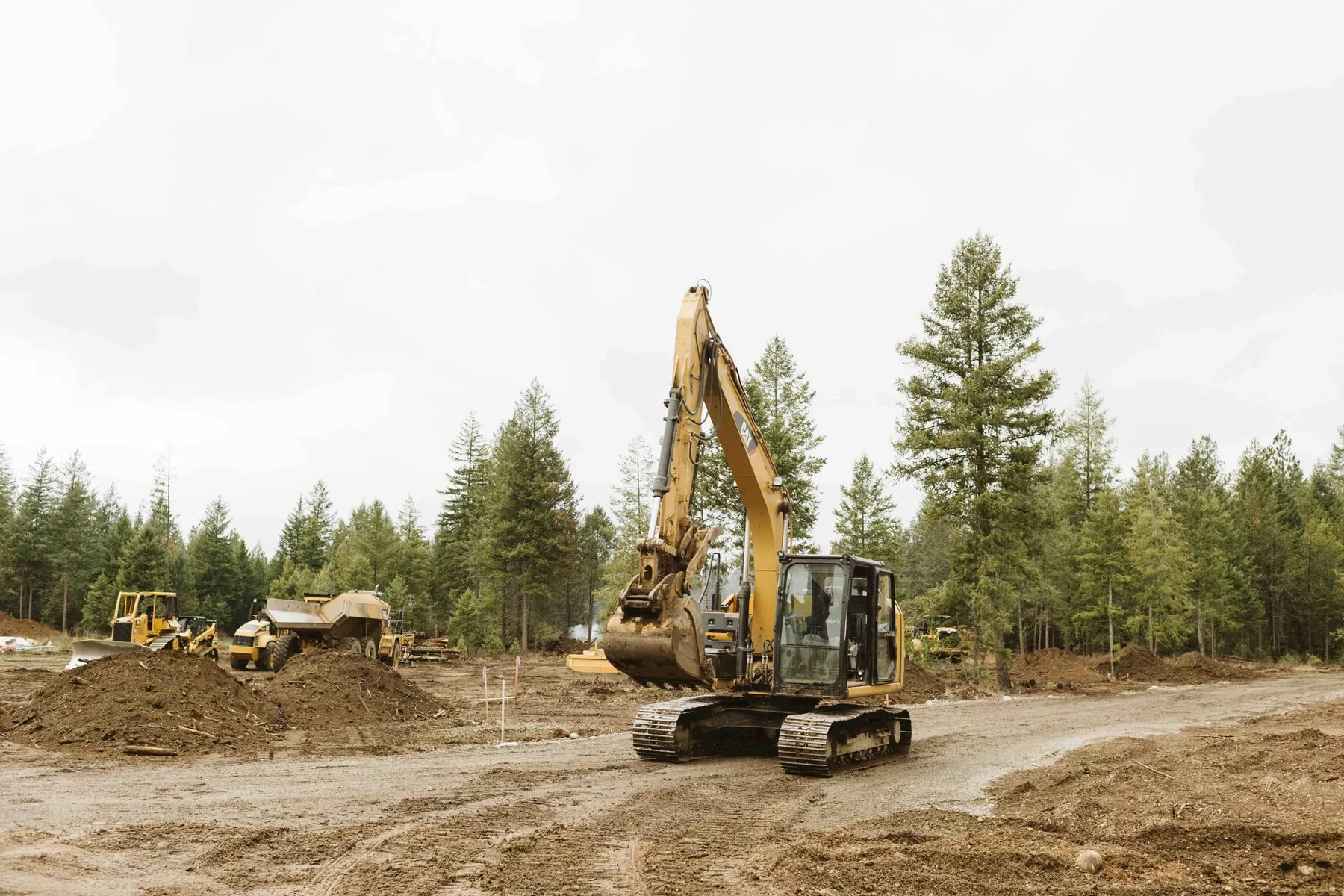 CAT excavator grading a cleared homesite, with a dozer and dump truck working in the background.