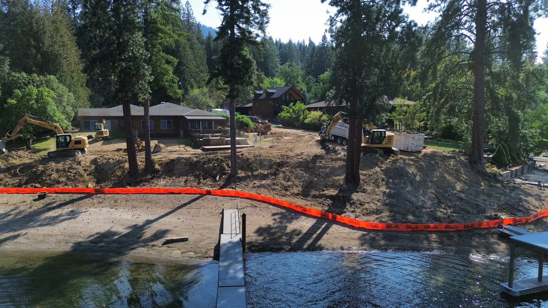 Lake-side view of the site during work, with an orange turbidity curtain in the water and excavators on the bank.