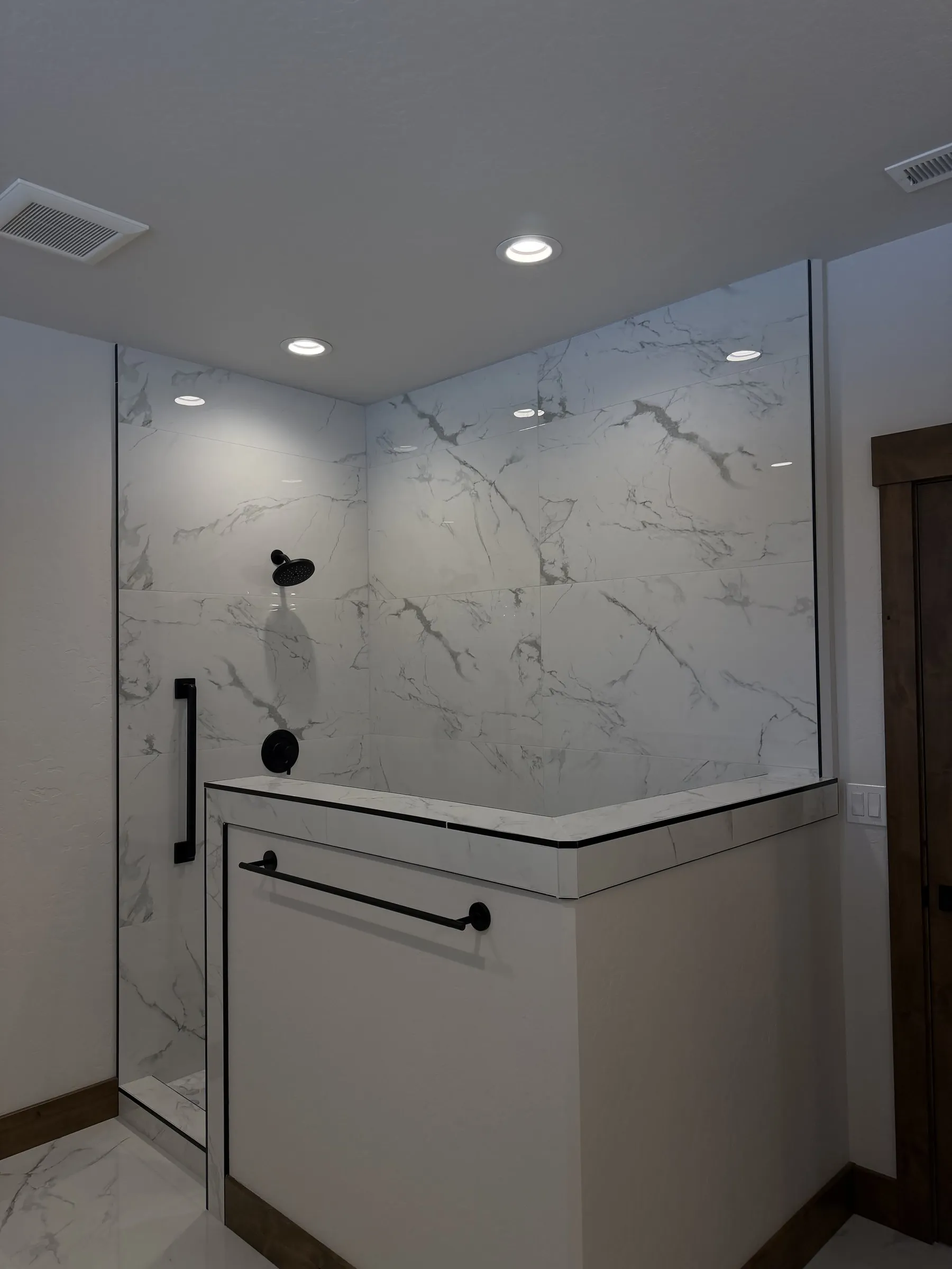 Walk-in shower with marble-look tile, a glass panel and black fixtures beside a half wall.