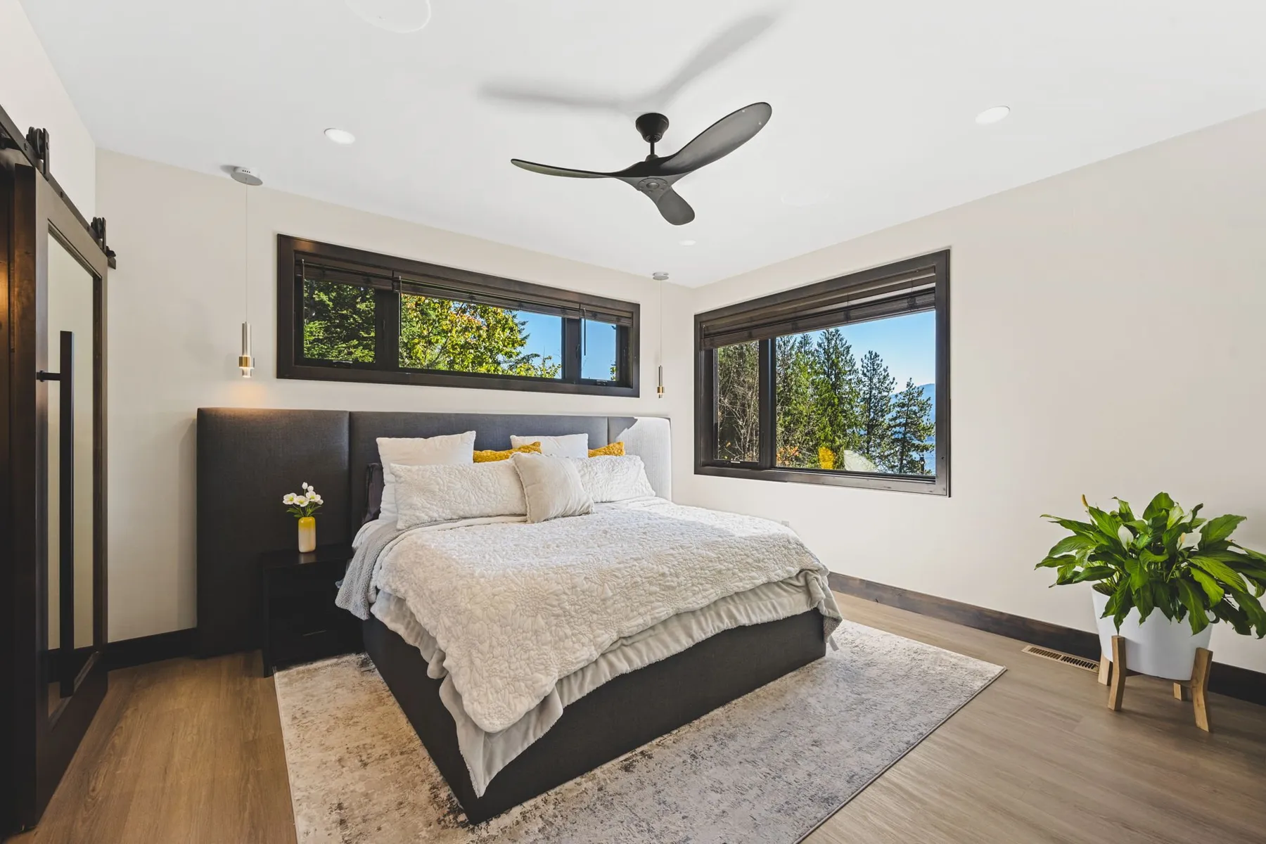 Primary bedroom with a black upholstered bed, ceiling fan and black-framed windows facing the lake and trees.