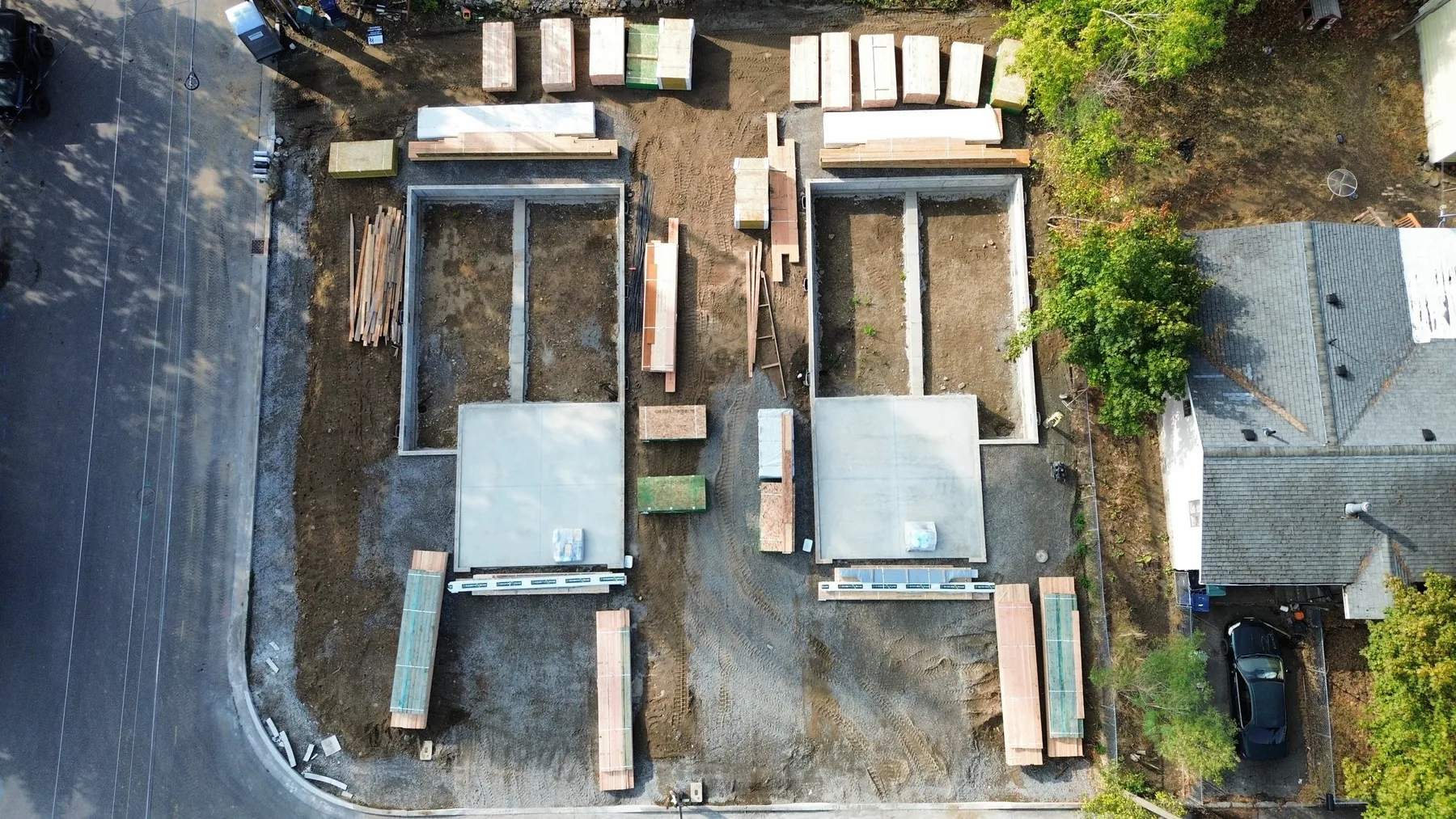 Aerial view of the twin townhome lot during construction, with two poured foundations and stacked framing lumber beside the street.