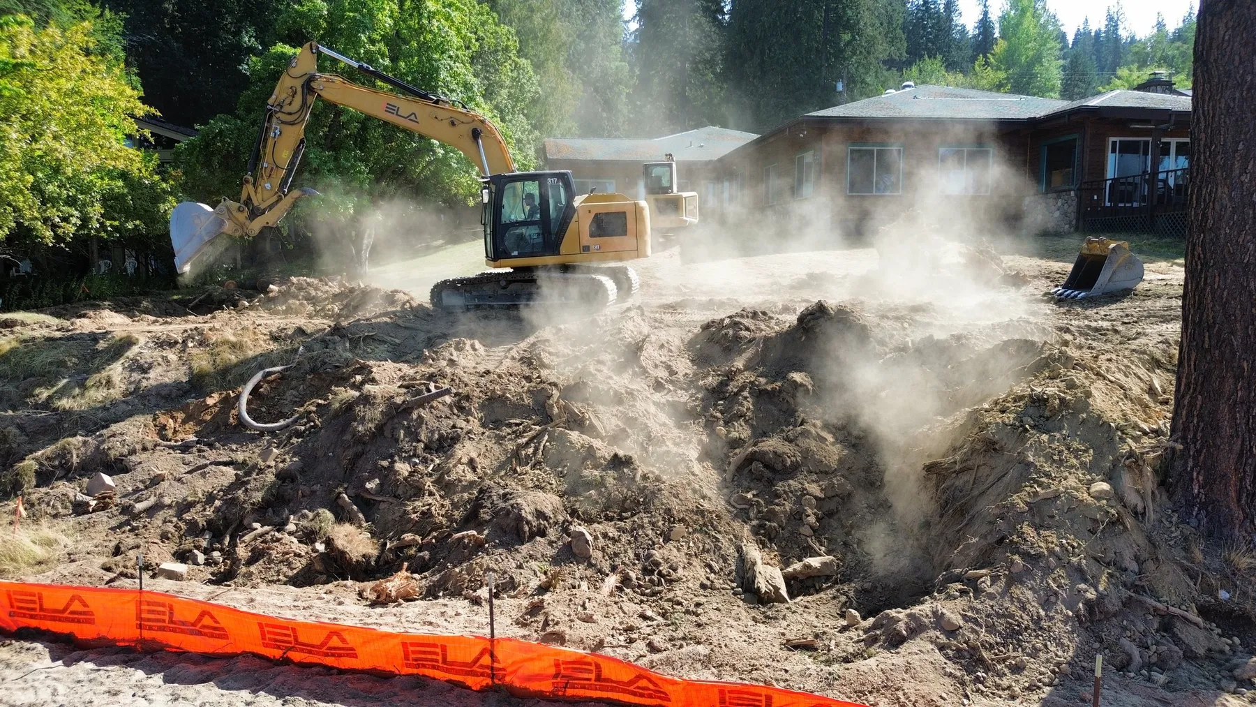 Excavator breaking up dusty ground behind a branded orange silt fence near a lakeside home.
