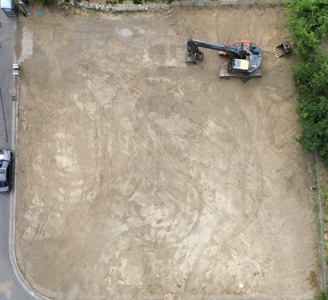 Aerial view of the cleared, graded corner lot before construction, with an excavator parked in one corner.