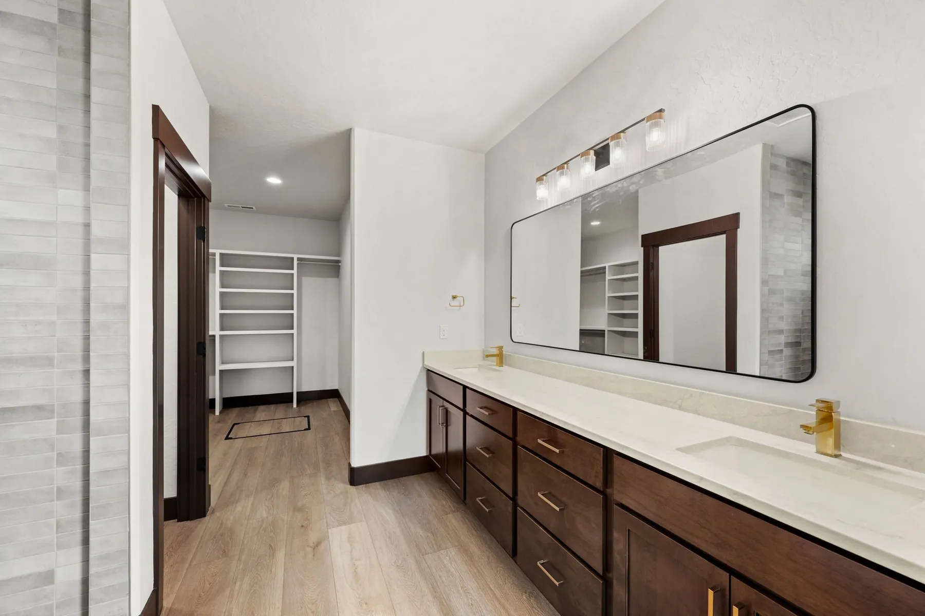 Primary bathroom with a dark wood dual vanity, gold fixtures and a large black-framed mirror, with the walk-in closet through the doorway.
