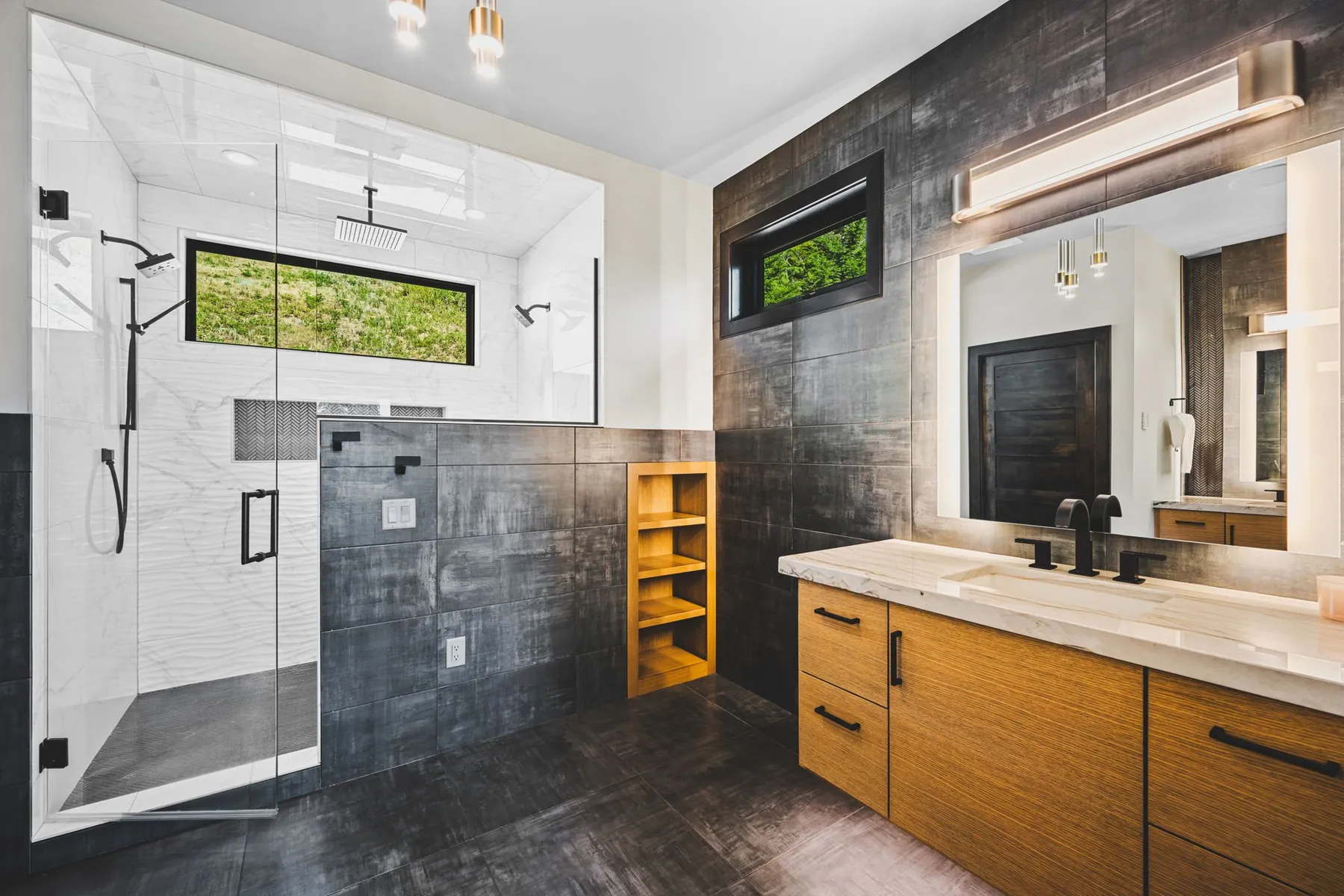 Spa-style primary bathroom with a glass walk-in shower, dark slate wall tile and a wood vanity with a lit mirror.