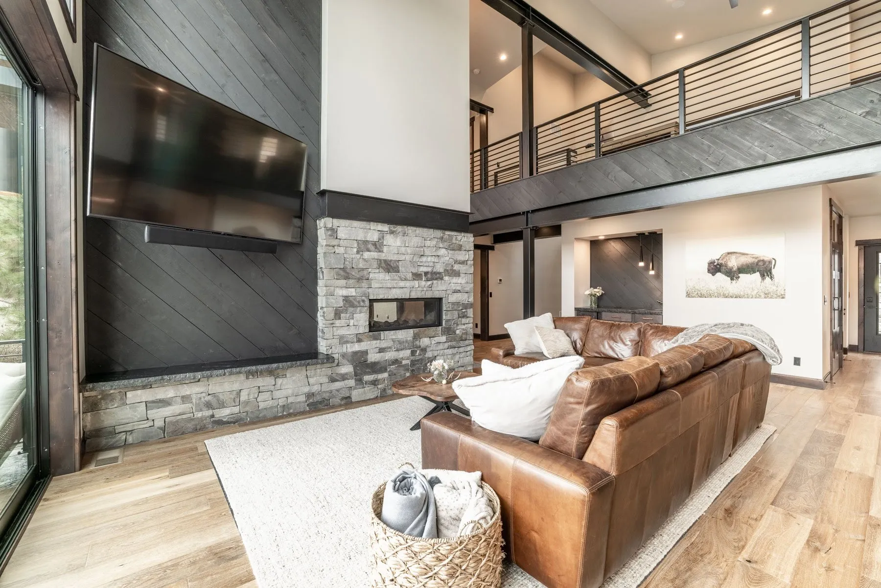 Lower family room with a leather sofa, a stacked-stone fireplace wall and an open catwalk railing above.