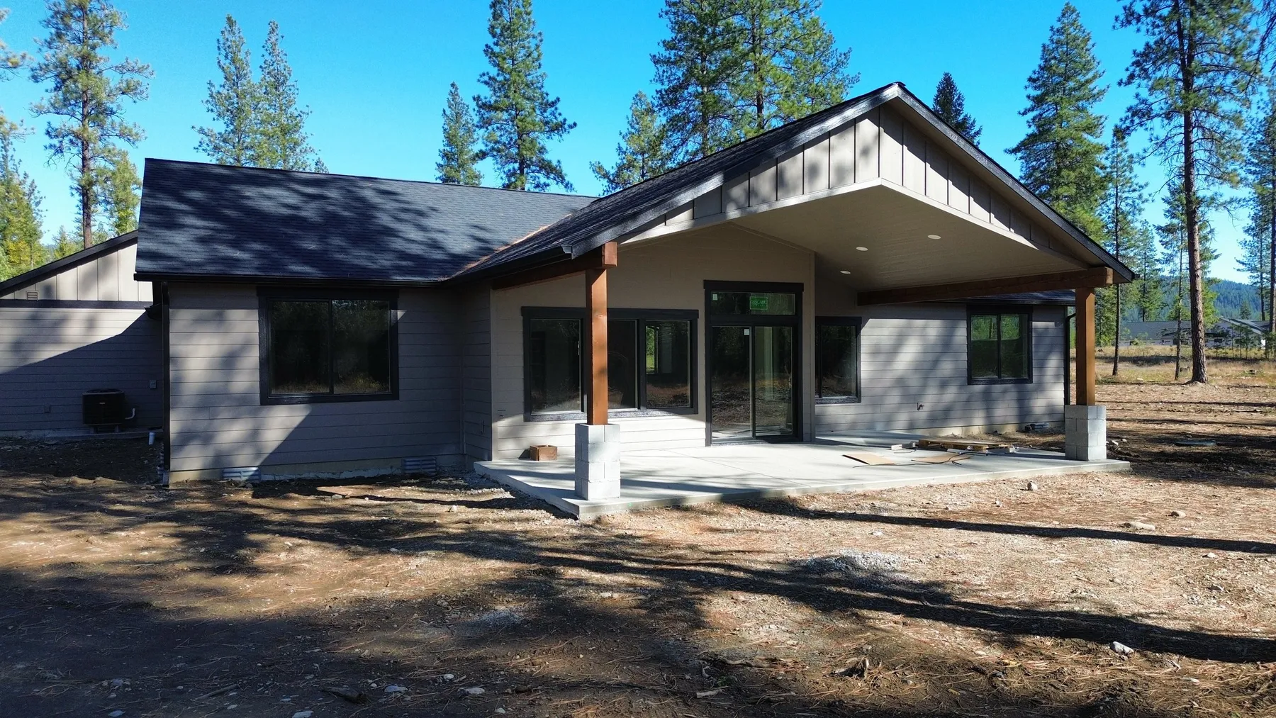 Back of the home with a gabled covered patio on timber posts and a concrete slab, surrounded by pines.