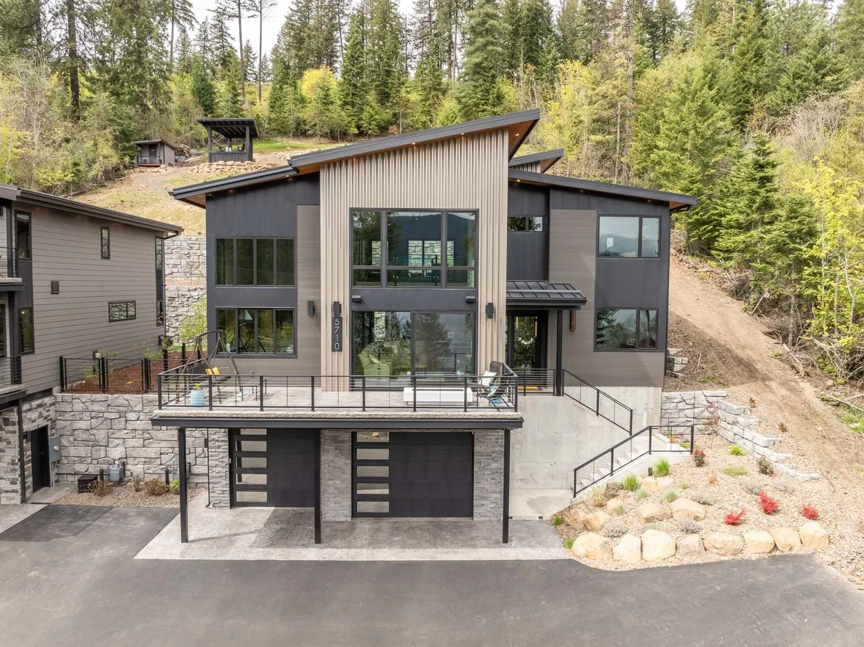 Three-story modern lakefront home with gray and wood-tone siding, a shed roof and a large front deck above the garage, set into a wooded hillside with stone retaining walls.