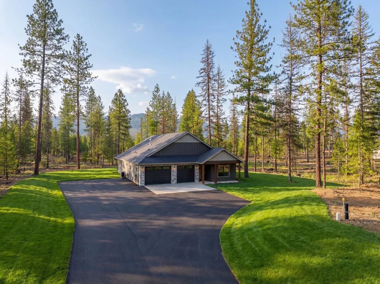 The Radiant, a single-story gray home with a two-car garage at the end of a new paved driveway, set on a green lawn among tall ponderosa pines.