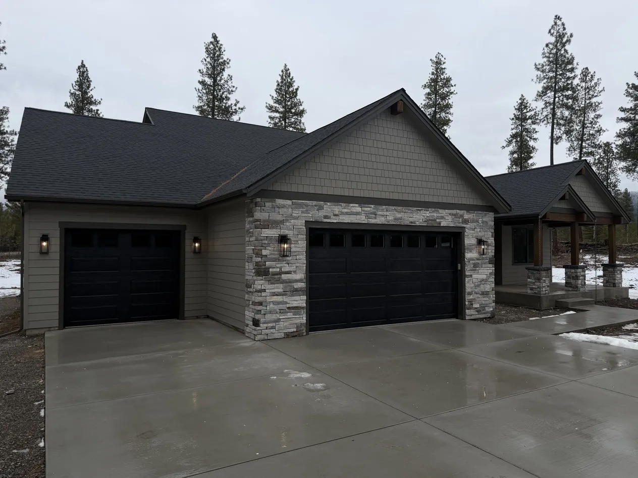 The Outlook, a gray craftsman home with a stacked-stone front, two black garage doors and a covered timber entry on a wet concrete driveway.