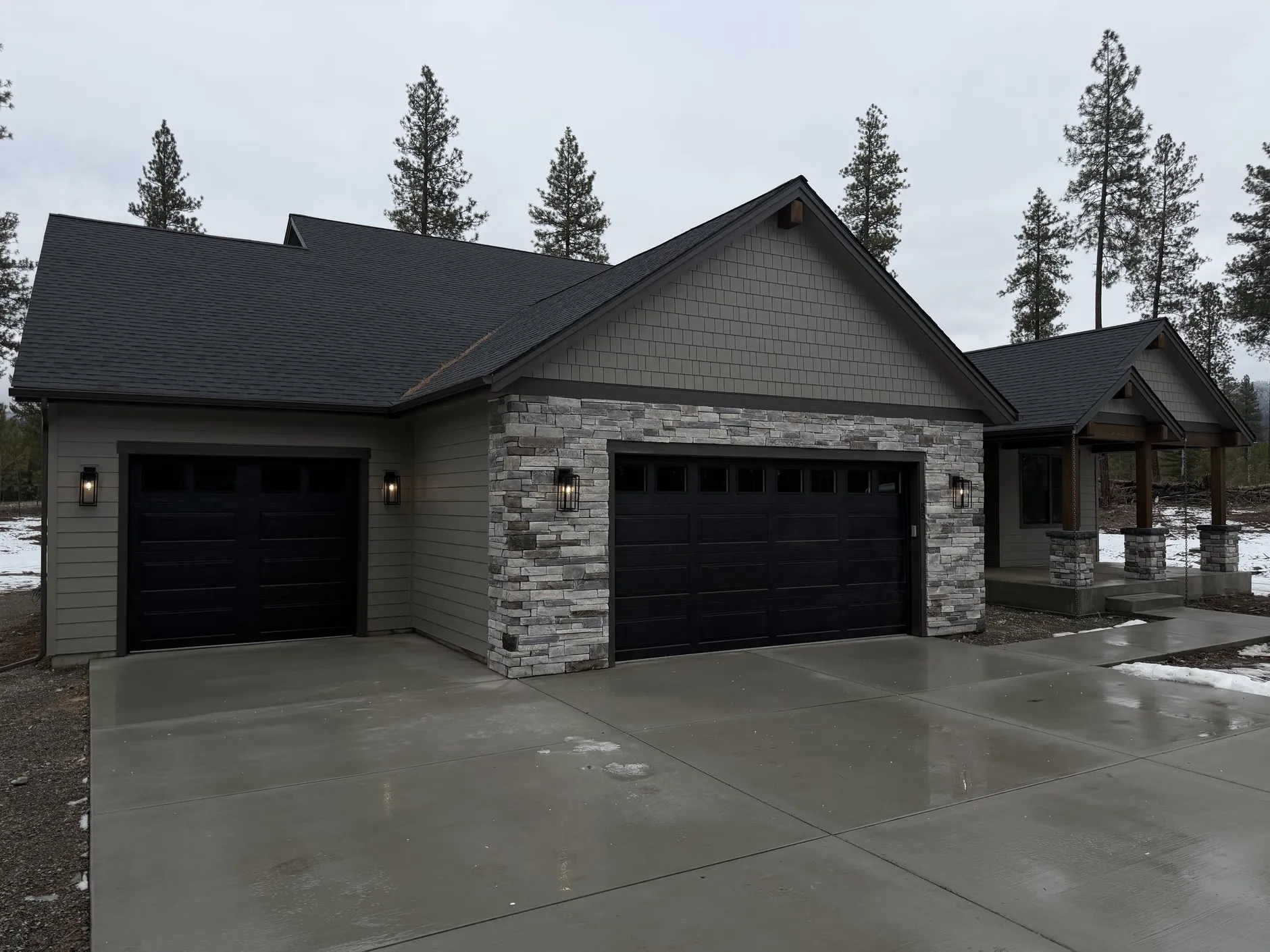 The Outlook, a gray craftsman home with a stacked-stone front, two black garage doors and a covered timber entry on a wet concrete driveway.