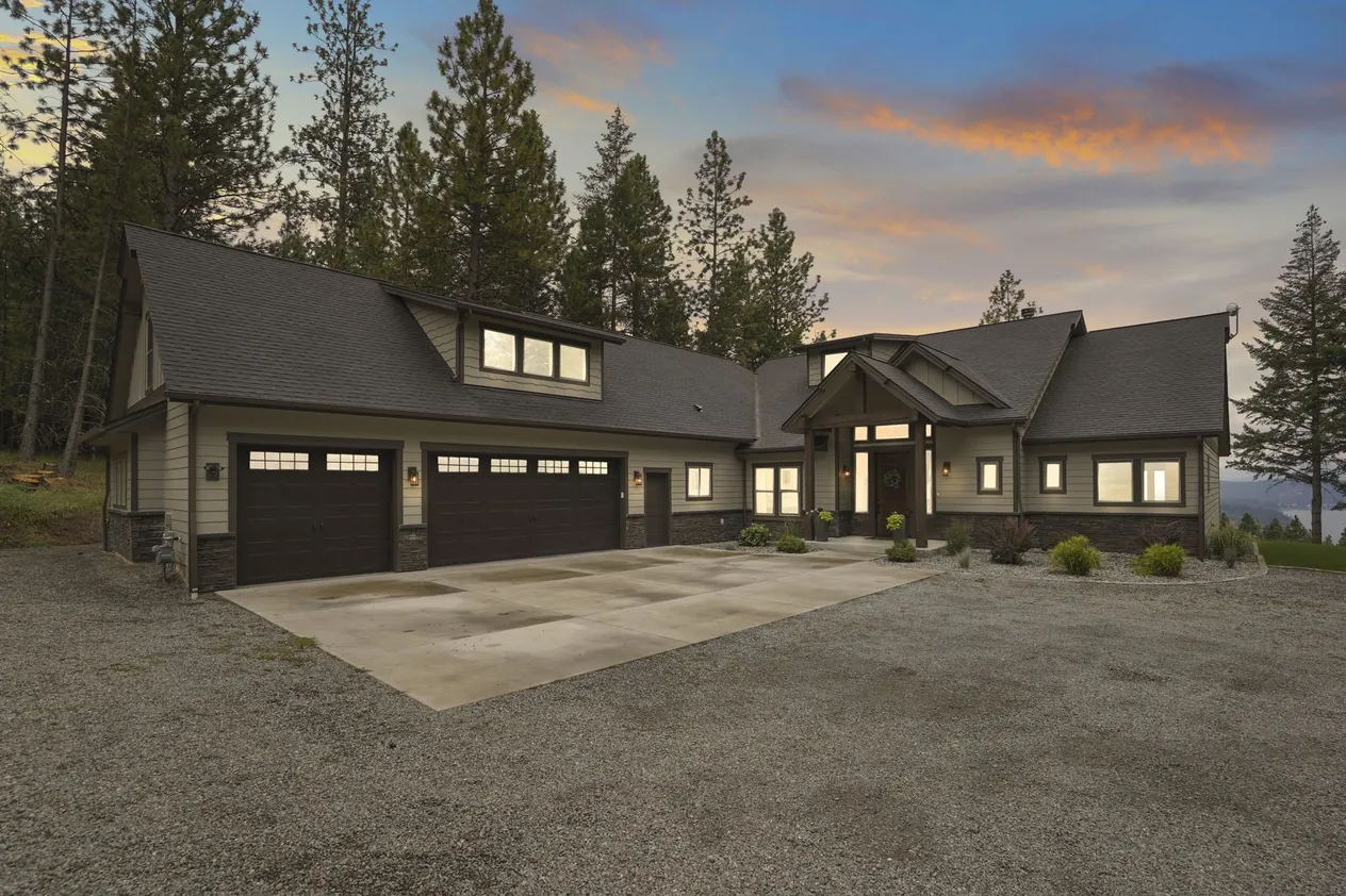 Lakeview Escape at dusk, a dark gray craftsman home with a three-car garage, lit windows and tall pines against a pink sky.