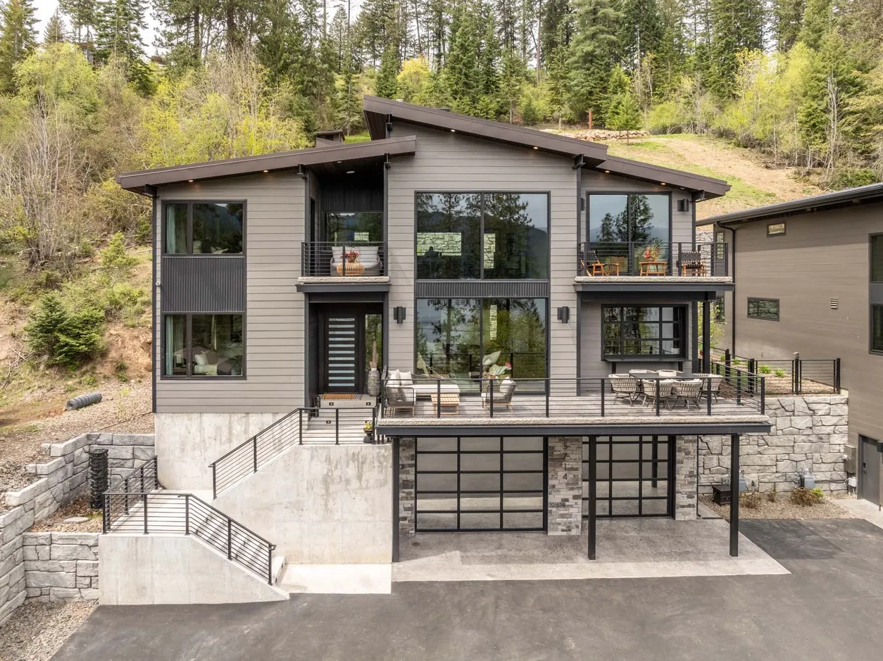 Three-story modern estate home on a wooded Hayden Lake hillside with railed decks on each level, a concrete estate wall and a glass garage door.