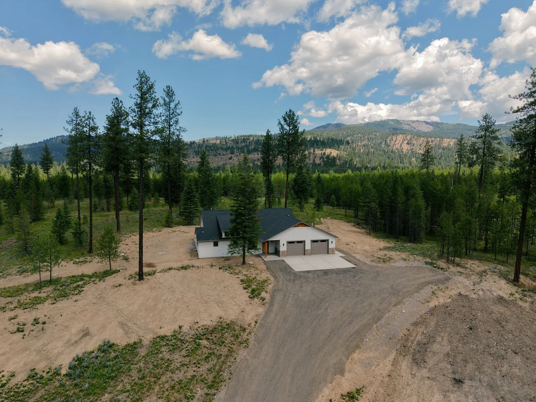 Aerial view of 93 Cannon, a new white farmhouse-style home on a cleared five-acre lot with a gravel drive, surrounded by pine forest and mountains.