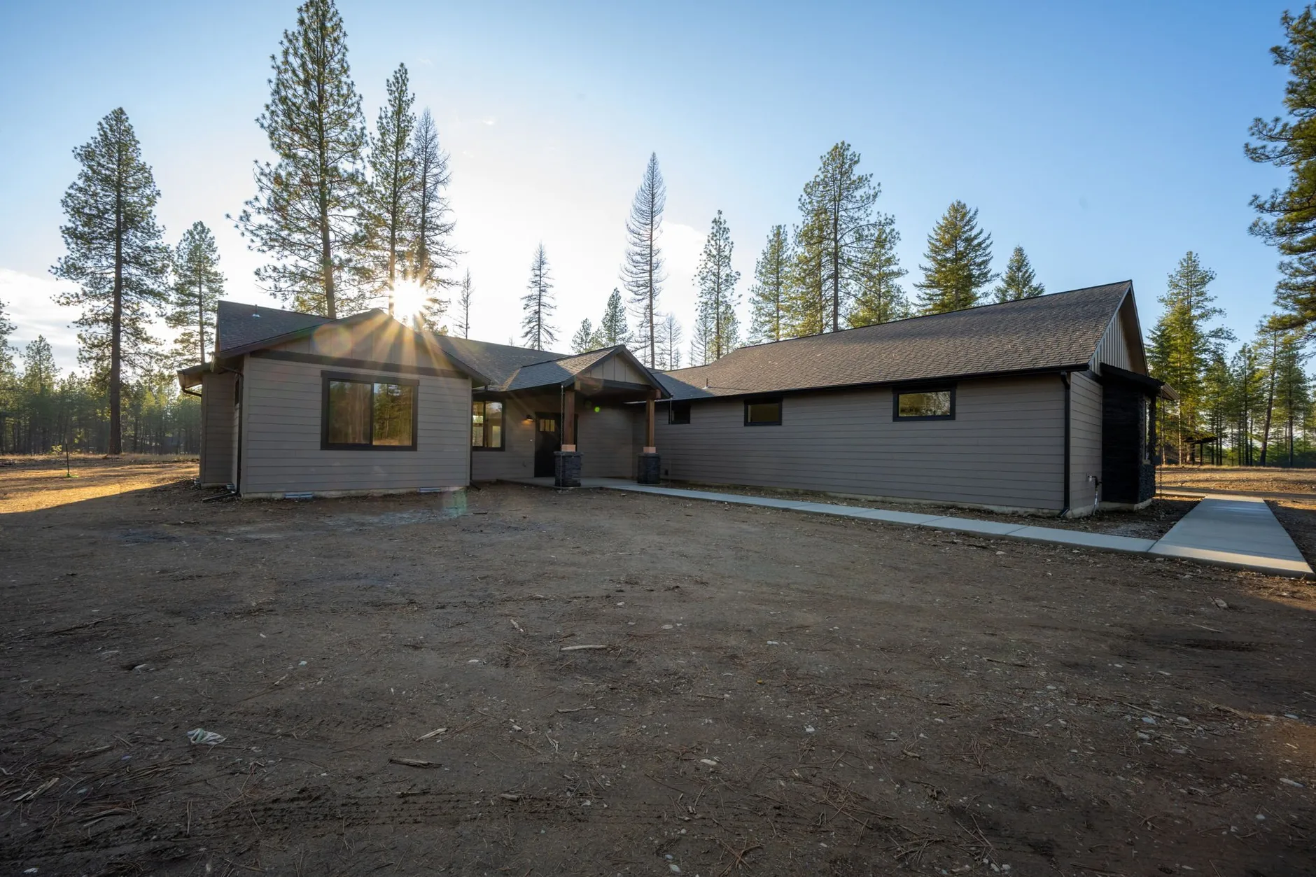 Single-story gray home with a covered entry on a freshly graded lot among tall pines, with the sun low behind the trees.