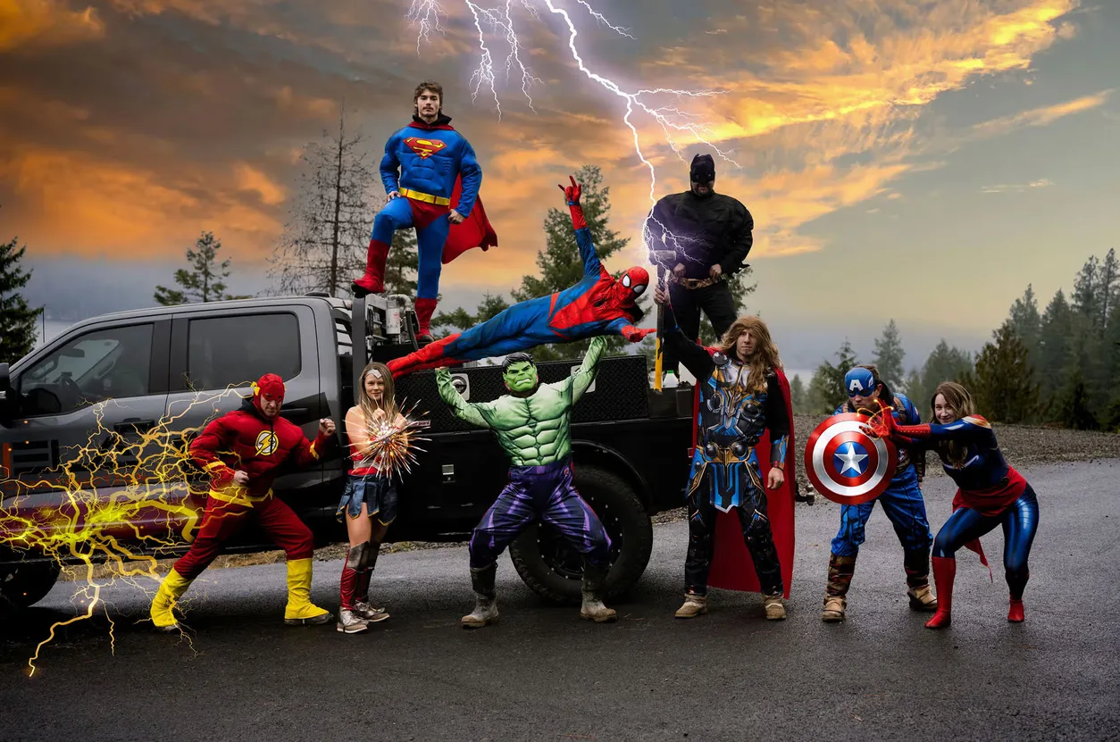 The Ela Construction crew dressed as superheroes (Superman, Batman, The Flash, Wonder Woman, Hulk, Spider-Man, Thor, Captain America and Captain Marvel) posing around a black Ela pickup truck under a stormy sunset sky with lightning.