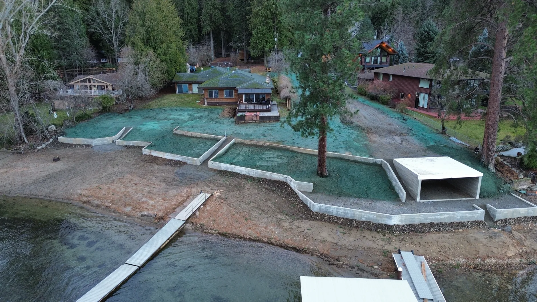 Aerial view of a Hayden Lake waterfront lot with new concrete seawalls, terraced hydroseeded beds and a flat-roofed concrete cabana built into the retaining walls beside a dock.