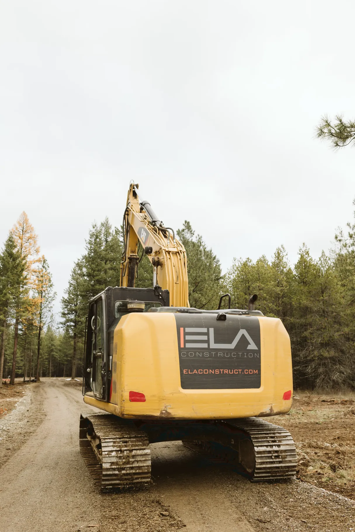 A CAT excavator on a dirt road among pine trees with the back facing us, and a black Ela Construction logo on it.