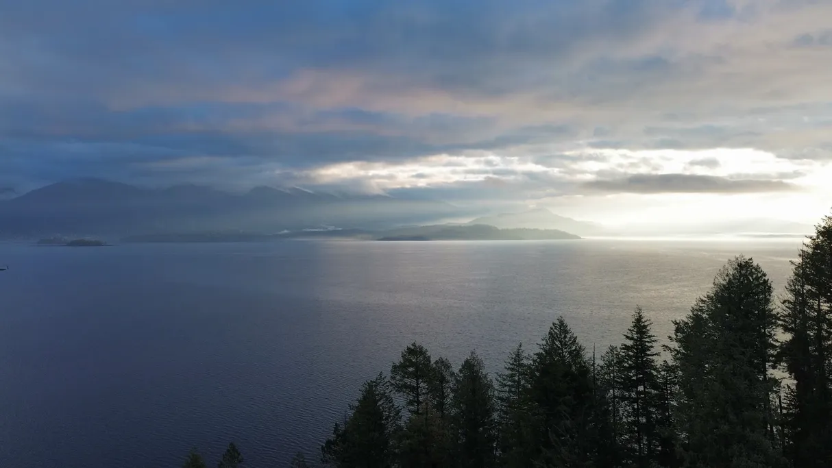 Aerial view of a wide North Idaho lake at sunrise, with low cloud over the mountains and dark evergreens in the foreground.