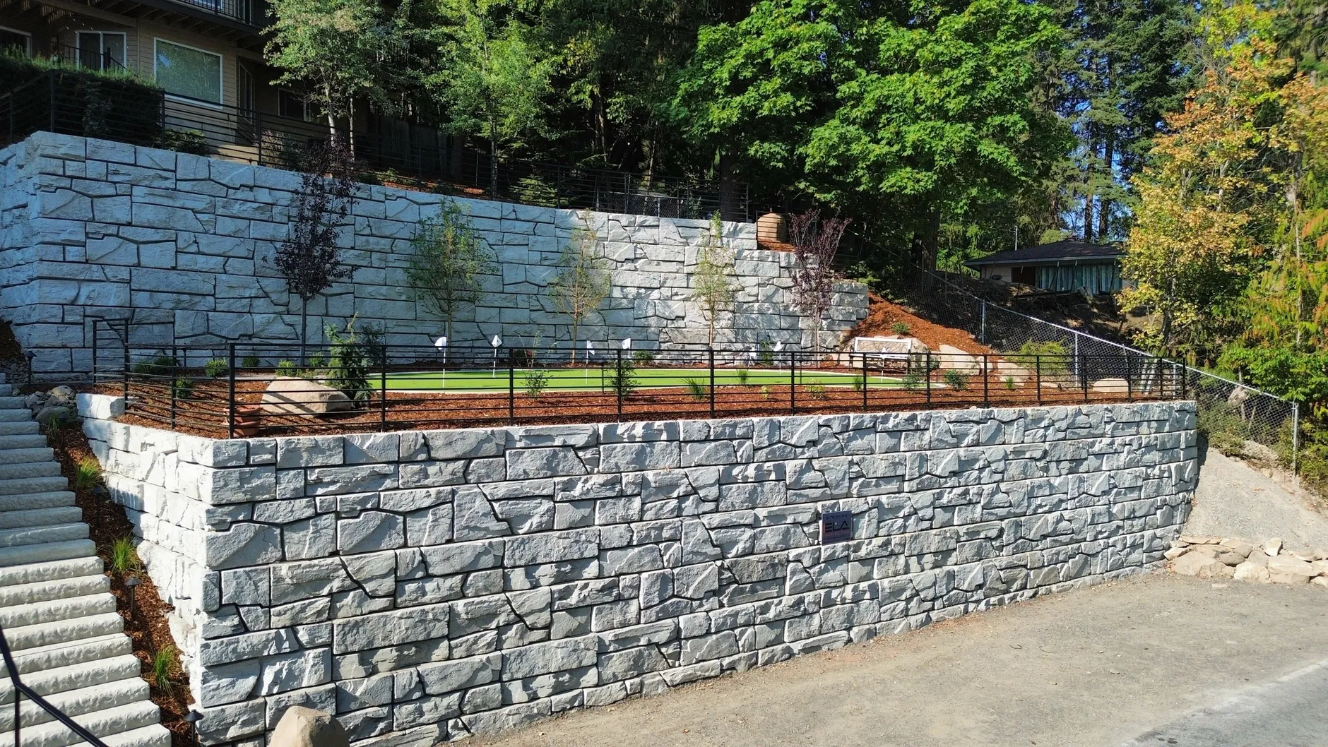 Two tiers of grey stone-textured retaining walls along Hayden Lake Road, with a turf terrace, black metal railing and stone steps between them.