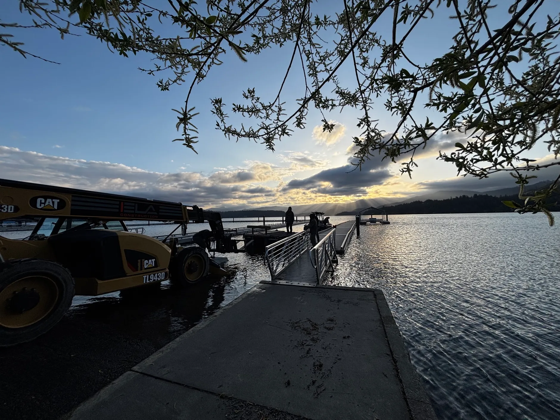 A CAT telehandler at a lakeside launch beside a new aluminum gangway and dock at sunset, with two workers out on the dock.