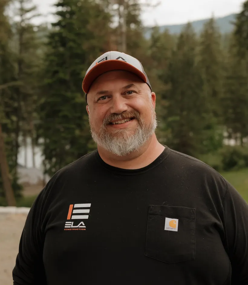 Anthony of Ela Marine, the marine division of Ela Construction, in a red-brimmed cap and black Ela T-shirt with evergreen forest behind him.
