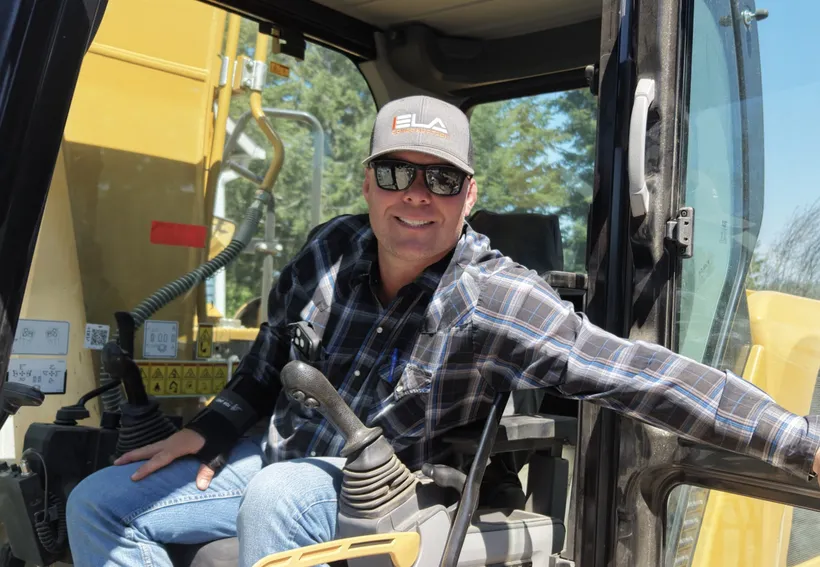 Derek, Director of Site Work Operations at Ela Construction, in sunglasses and an Ela cap, sitting in the open cab of a yellow excavator.