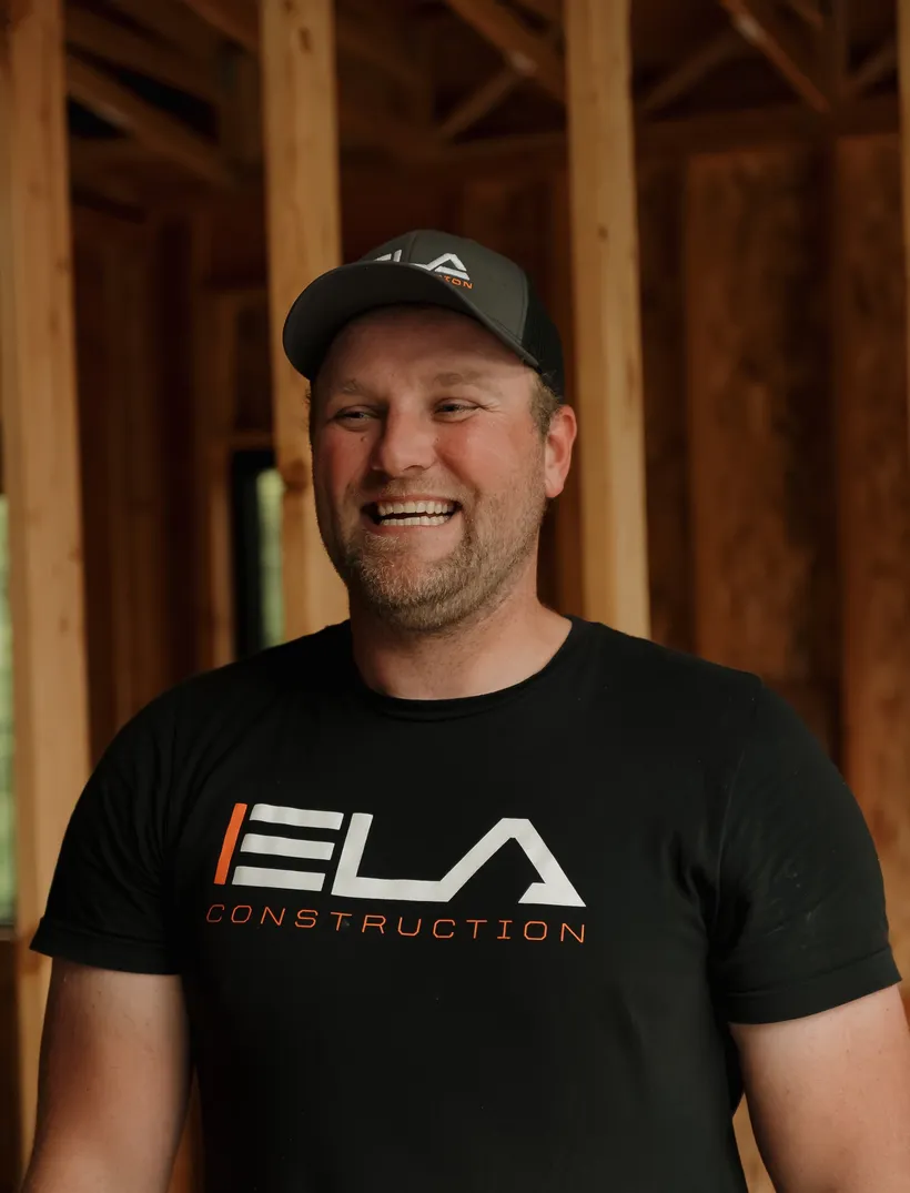 Dustin, Superintendent at Ela Construction, in a black Ela Construction T-shirt inside the timber frame of a house under construction.