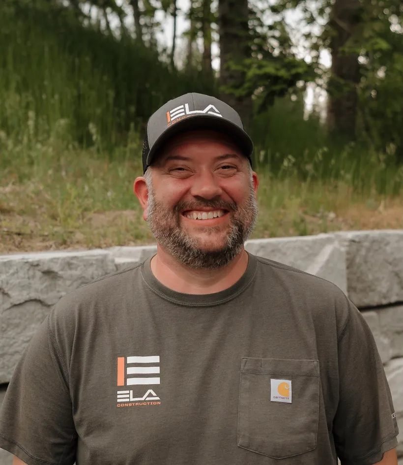 Garhard (G), Field Operations at Ela Construction, smiling in an Ela cap and olive Ela T-shirt in front of a stone retaining wall.