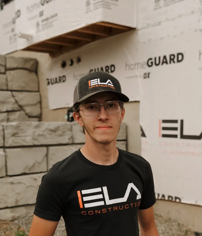 Garrett, Laborer at Ela Construction, in glasses, an Ela cap and a black Ela Construction T-shirt in front of a block wall and a wrapped house.
