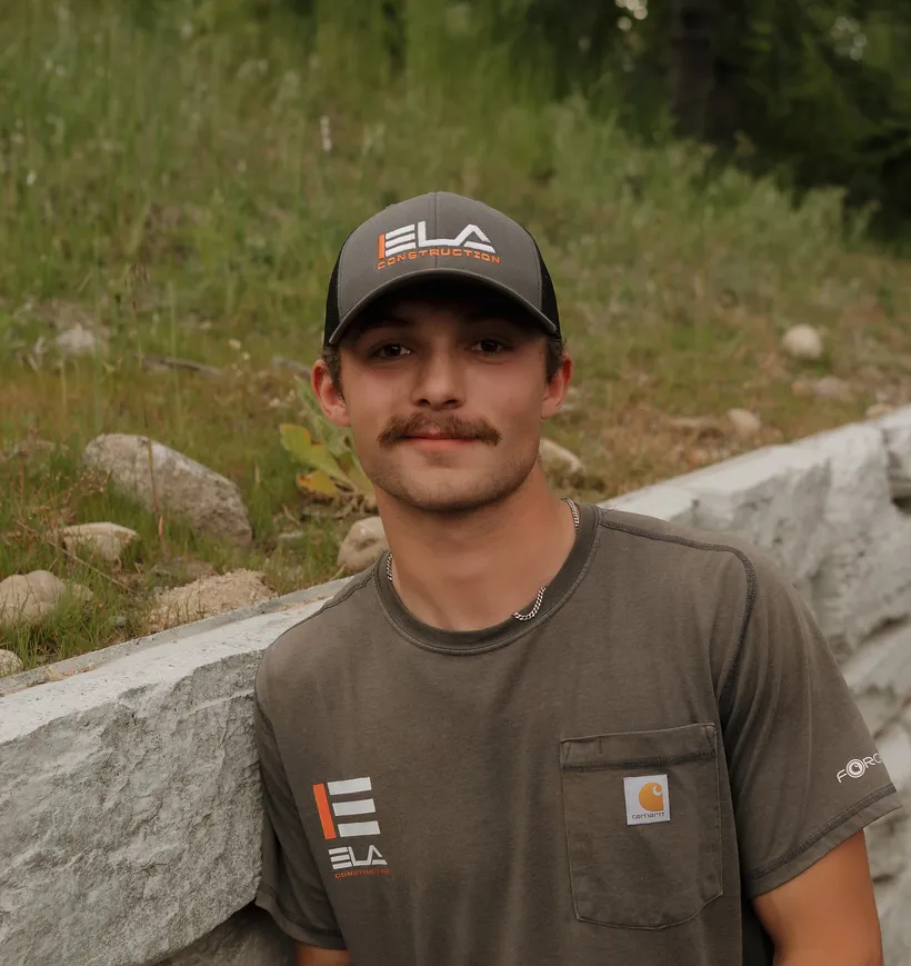 Jesse, Operator at Ela Construction, in an Ela cap and grey Ela T-shirt leaning against a stone block retaining wall.