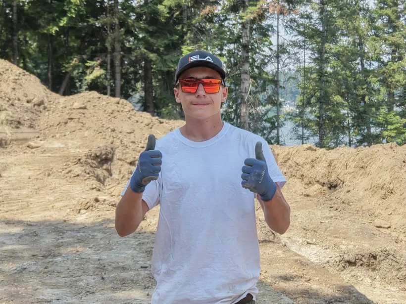 Kaleb, Laborer at Ela Construction, giving two thumbs up in orange safety glasses and work gloves on an excavated dirt site.