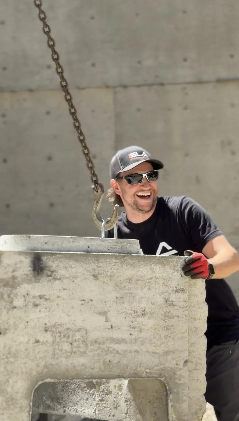 Luke, Truck Driver at Ela Construction, in sunglasses and an Ela cap guiding a concrete block hanging from a lifting chain.