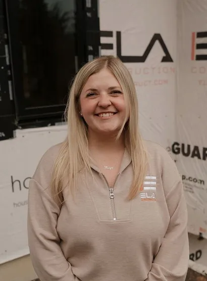 Megan, Financial Operations at Ela Construction, smiling in a beige Ela quarter-zip in front of a house wrapped in building paper.