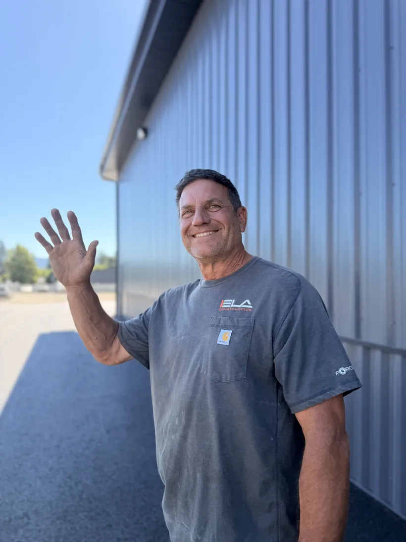Mike, Equipment Mechanic at Ela Construction, waving beside a blue metal shop building on a sunny day.