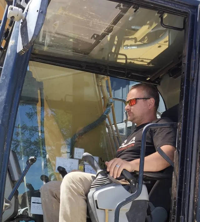 Patrick, Operator at Ela Construction, in sunglasses and an Ela T-shirt at the controls inside an excavator cab.