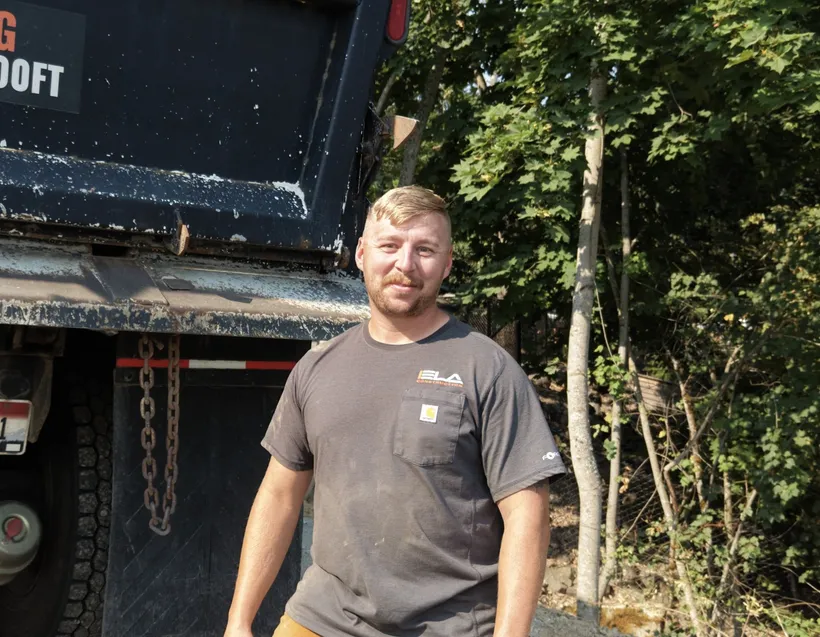 Randall, Truck Driver at Ela Construction, standing at the back of a dump truck at the edge of a wooded lot.