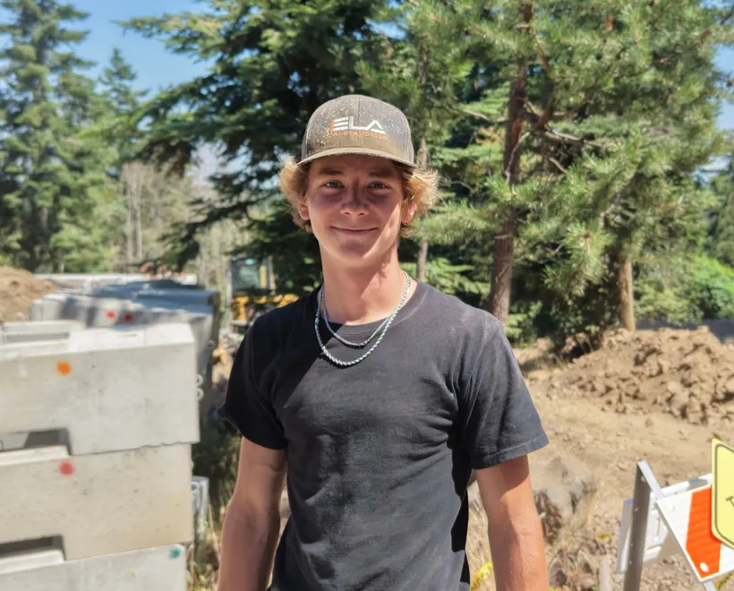 Sawyer, Laborer at Ela Construction, in an Ela cap and black T-shirt beside stacked concrete blocks on a dirt job site among pine trees.